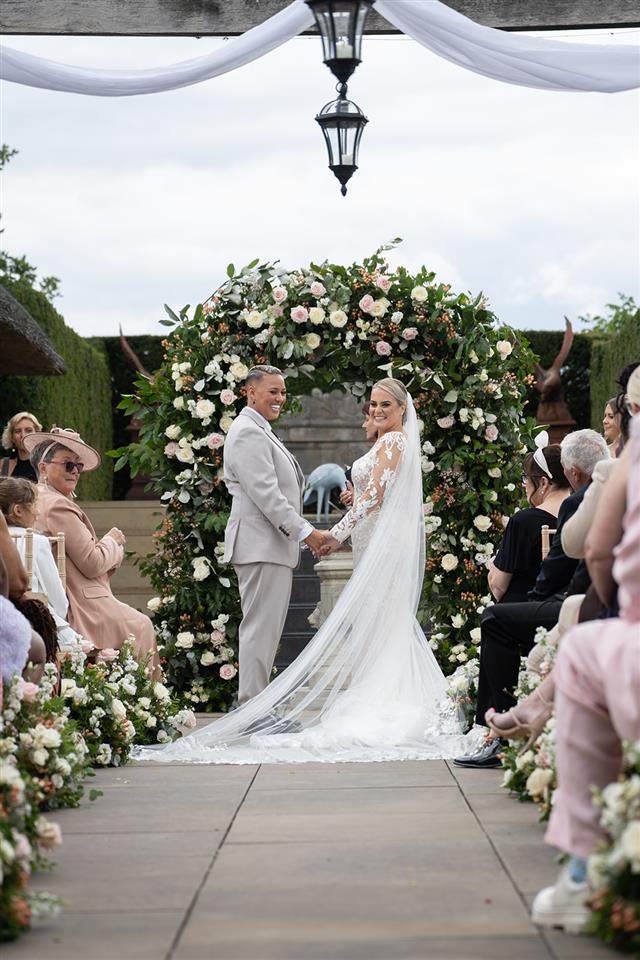 Outdoor wedding ceremony taking place with flower meadows down the aisle and a large floral arch
